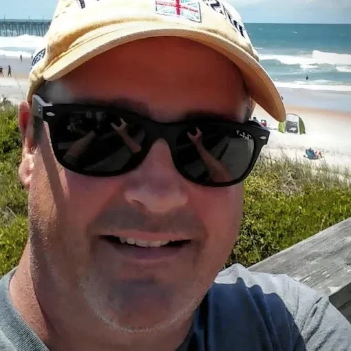 A man wearing sunglasses smiling for a selfie at the beach.
